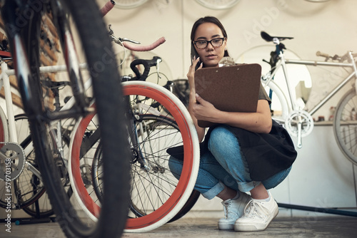 Is it the white or pink one. Full length shot of an attractive young woman crouching in her bicycle shop and using her cellphone while holding a clipboard.