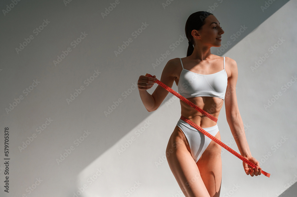 Red colored measuring tape in hands. Young caucasian woman with slim body shape is indoors in the studio Red colored measuring tape in hands. Young caucasian woman with slim body shape is indoors in the studio