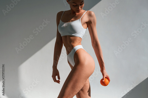 With an orange in hand. Young caucasian woman with slim body shape is indoors in the studio