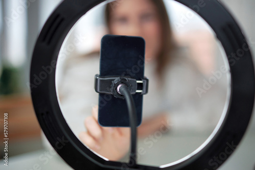 Lifestyle beauty blogger streaming online from home studio. Diverse millennial woman working as make up artist and recording video for social media on smartphone with led ring lamp, blogging content 