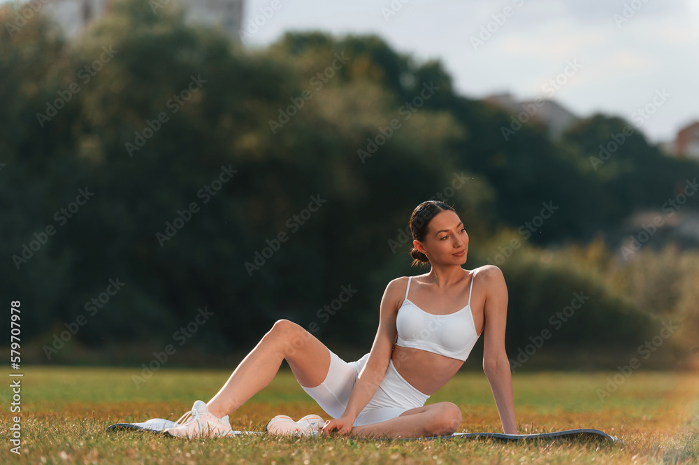 Enjoying nature. Sitting on the mat. Young caucasian woman with slim body shape is in the fitness clothes outdoors