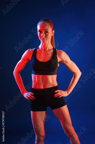 Standing with the hands on waist. Beautiful muscular woman is indoors in the studio with neon lighting