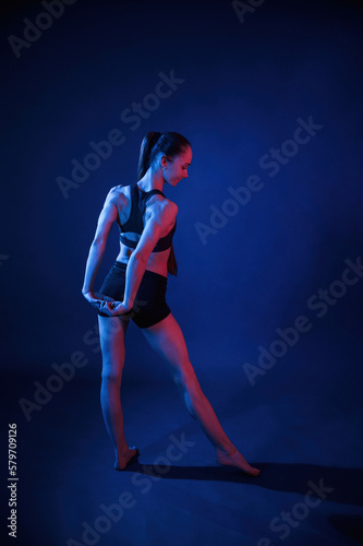 Back view. Warm up stretching exercises. Beautiful muscular woman is indoors in the studio with neon lighting