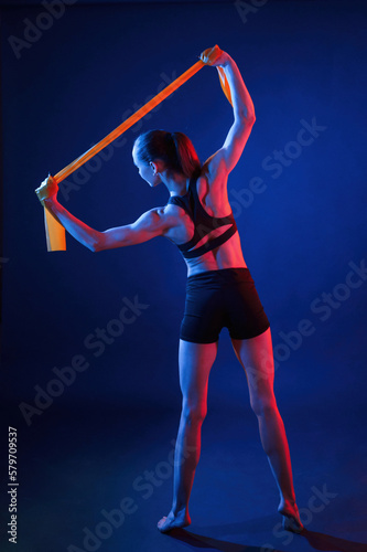 Yellow colored tape for stretching the hands. Beautiful muscular woman is indoors in the studio with neon lighting