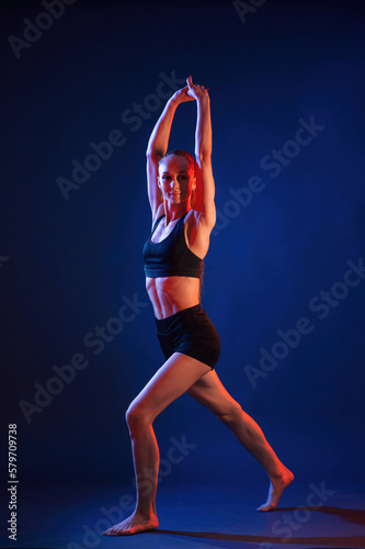 Sportive exercises, standing and stretching the hands. Beautiful muscular woman is indoors in the studio with neon lighting