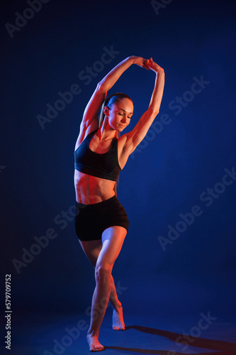 Sportive exercises, standing and stretching the hands. Beautiful muscular woman is indoors in the studio with neon lighting