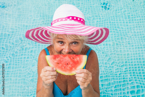 Happy senior woman eating juicy watermelon slice at the swimming pool during summertime photo