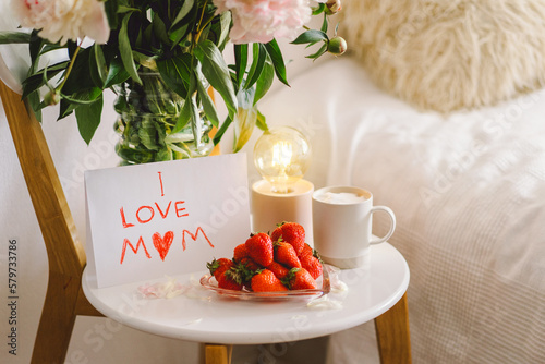 Breakfast for Mothers Day. Heart shaped white plate with fresh strawberries, cup of coffee, gift and Peonys bouquet with gift in bed. Still life composition. Happy Mother's Day.