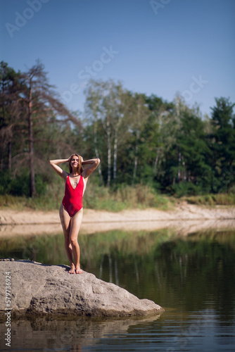 A young girl with long blond hair  with a slender figure  in a red bathing suit  stant on a stone  in a river  against the backdrop of a forest.