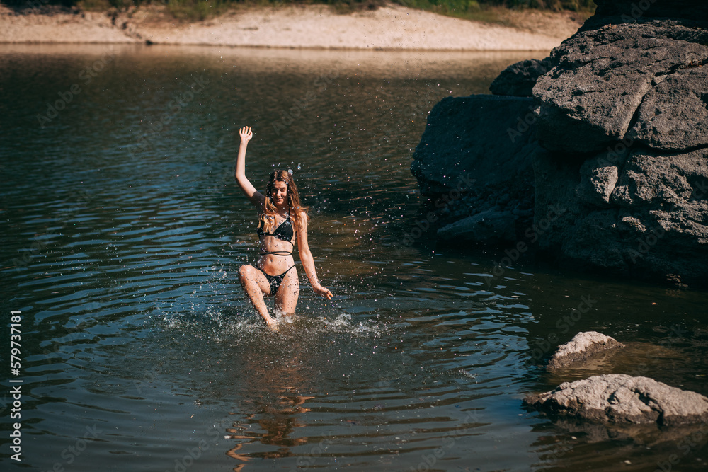 Fototapeta premium A young girl with long blond hair, a slender figure, in a black swimsuit jumps in the river against the backdrop of rocks and laughs.