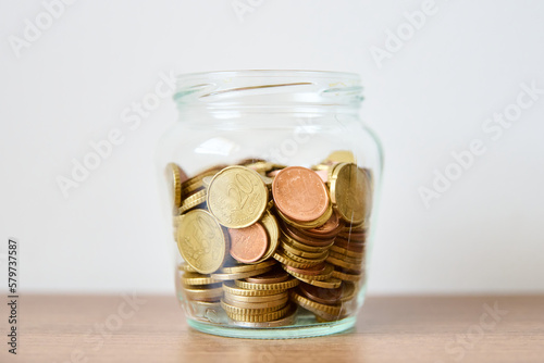 Coins in glass jar on wooden table and white wall background.