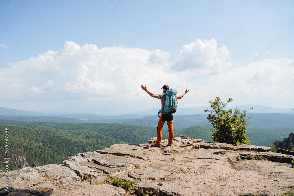 Obraz premium guy hiker standing on the edge of a cliff keeping his hands to the sides, victorious arm raise, big backpack on his back, mountain climbing, outdoor vacation