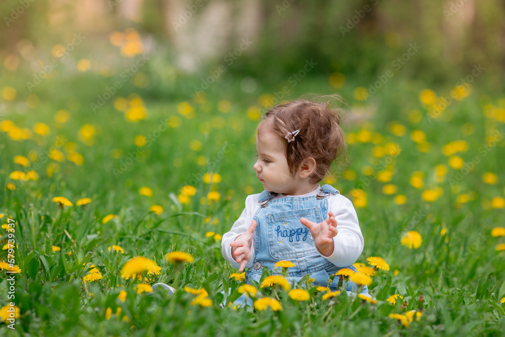 Happy little baby girl sitting on a green meadow with yellow flowers dandelions on the nature in the park Happy little baby girl sitting on a green meadow with yellow flowers dandelions on the nature in the park