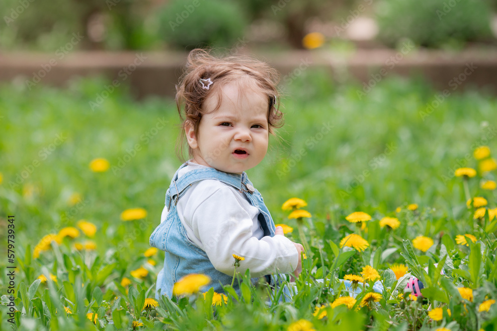 Happy little baby girl sitting on a green meadow with yellow flowers dandelions on the nature in the park Happy little baby girl sitting on a green meadow with yellow flowers dandelions on the nature in the park
