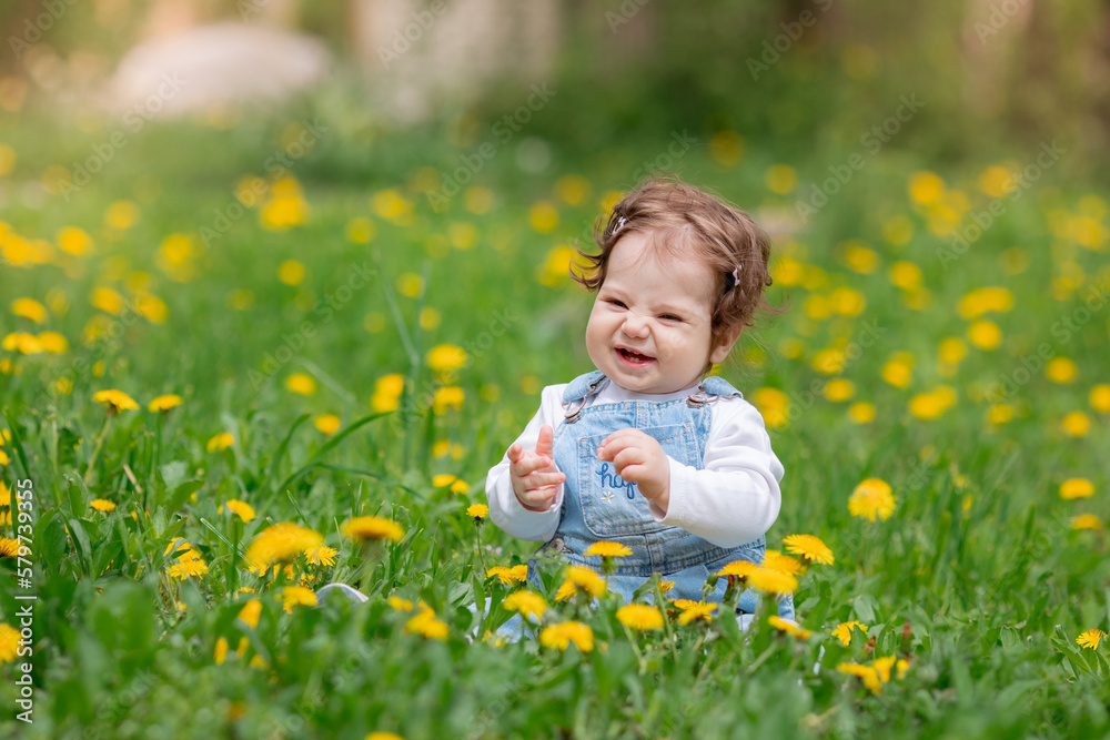 Happy little baby girl sitting on a green meadow with yellow flowers dandelions on the nature in the park Happy little baby girl sitting on a green meadow with yellow flowers dandelions on the nature in the park