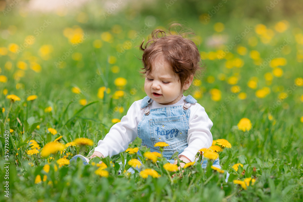 Happy little baby girl sitting on a green meadow with yellow flowers dandelions on the nature in the park Happy little baby girl sitting on a green meadow with yellow flowers dandelions on the nature in the park
