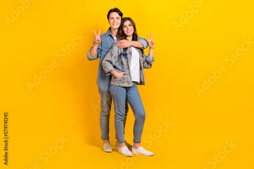 Full length photo of sweet pretty couple wear jeans showing v-signs cuddling isolated yellow color background