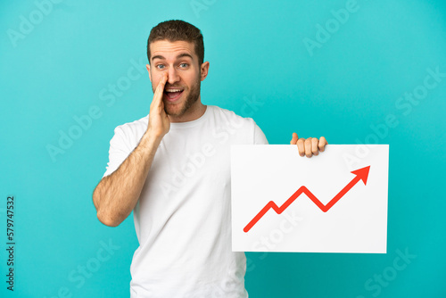 Handsome blonde man over isolated blue background holding a sign with a growing statistics arrow symbol and shouting