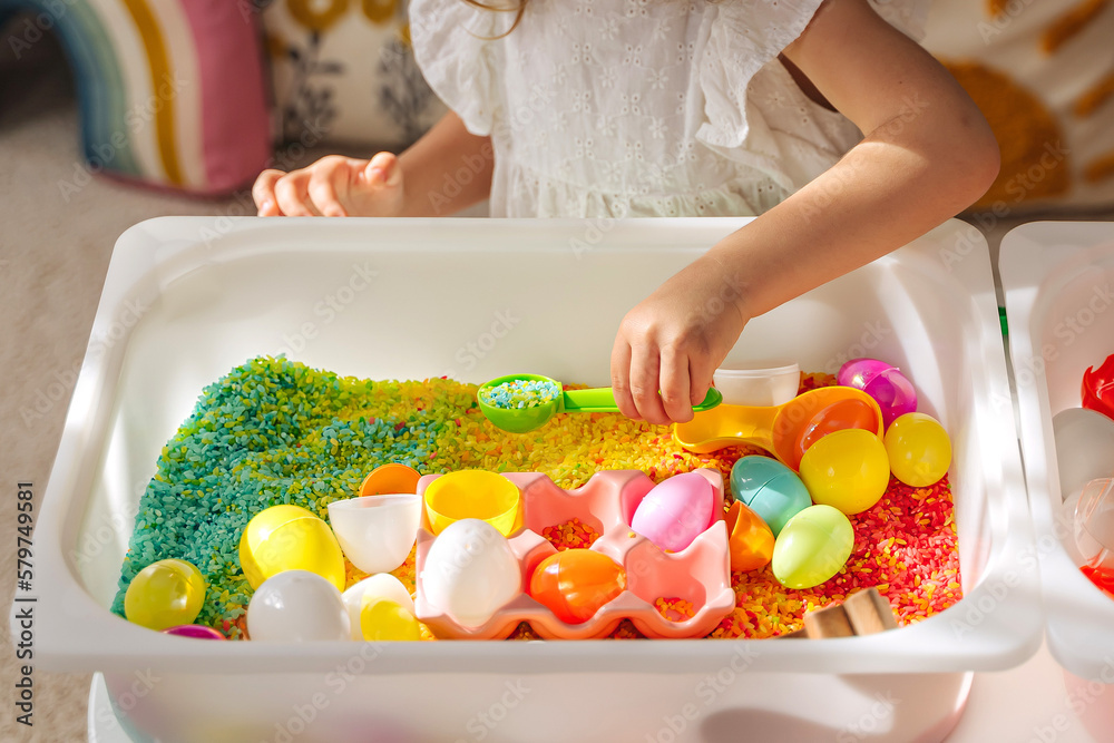 A little girl playing with colored rice and Easter eggs in sensory bin. Easter sensory bin with bright rice and eggs, bunny, carrot. Sensory play and holidays activity for kids. Happy Easter concept. - obrazy, fototapety, plakaty 