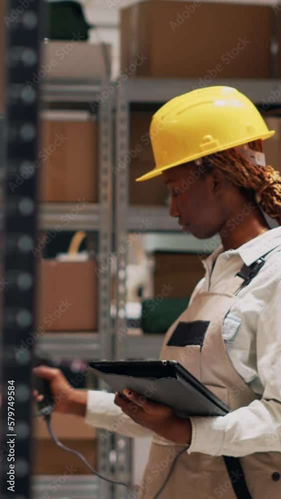 Vertical video: Young adult in overalls checking list of products on tablet, using scanner to count number of merchandise boxes and packages. Female employee working in storage room with goods. Vertical video: Young adult in overalls checking list of products on tablet, using scanner to count number of merchandise boxes and packages. Female employee working in storage room with goods.