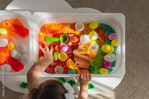 Child playing with colored rice and Easter eggs in sensory bin. Easter sensory bin with bright rice and eggs, bunny, carrot. Sensory play and holidays activity for kids. Happy Easter concept.