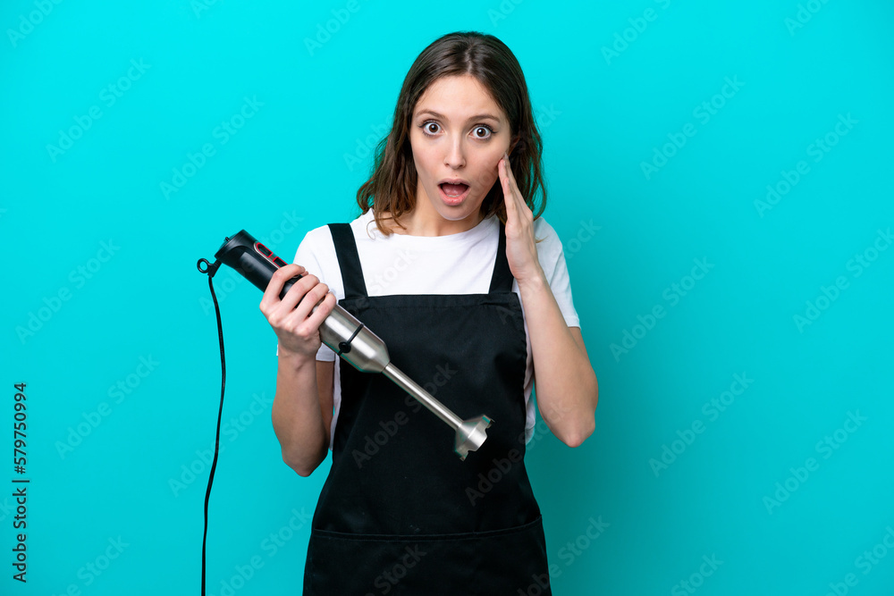 Young caucasian cooker woman using hand blender isolated on blue background with surprise and shocked facial expression