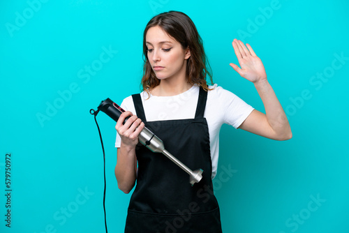 Young caucasian cooker woman using hand blender isolated on blue background making stop gesture and disappointed