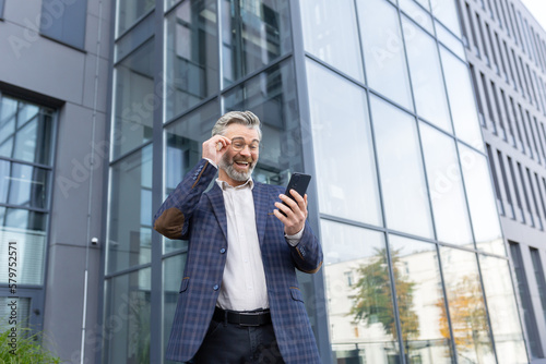 A happy senior gray-haired man in a suit and glasses stands outside an office, a skyscraper. Joyful reads a message on the phone, received good news, adjusts his glasses.