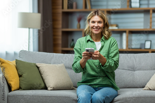 Photo of a senior woman housewife sitting on the sofa and using the phone, smiling at the camera.