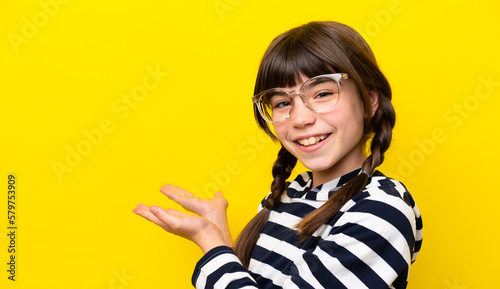 Little caucasian girl isolated on yellow background With glasses and presenting something