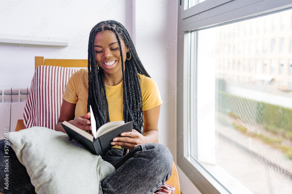 Smiling black woman reading book on armchair near window Smiling black woman reading book on armchair near window