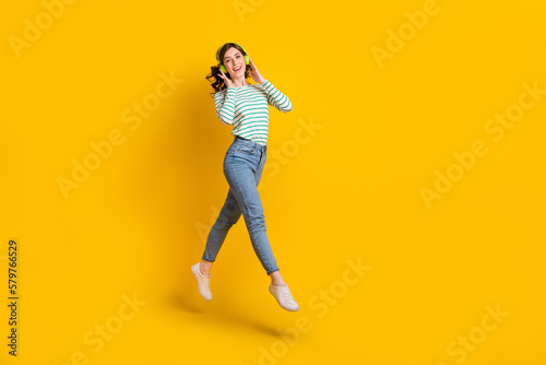 Full length photo of good mood nice girl wear striped shirt denim pants flying touching headphones isolated on yellow color background