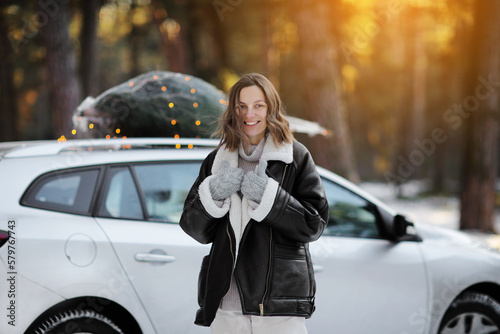 Smiling caucasian woman in winter coat and mittens near car with illuminated Christmas tree on a rooftop on nature in snowy forest. Concept of celebrating New Year holidays. Idea of Xmas mood and fun