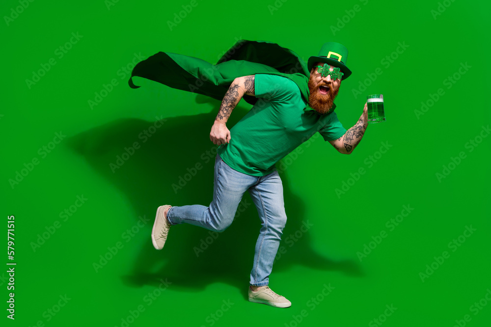 Full length photo of cool excited man wear t-shirt headwear drinking ale beer glass enjoying shamrock day isolated green color background Full length photo of cool excited man wear t-shirt headwear drinking ale beer glass enjoying shamrock day isolated green color background