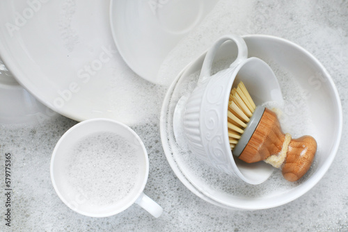 White dishes plates mugs in sink of dishwater with soap bubbles and scrub brush