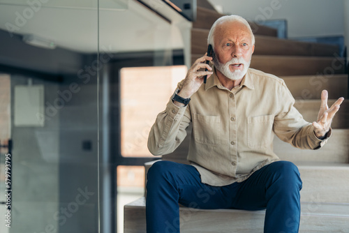 Senior man talking on mobile phone, looking upset and extremely nervous about hearing the bad news over phone call, sitting indoors on stairs, gesturing with hand and looking away.