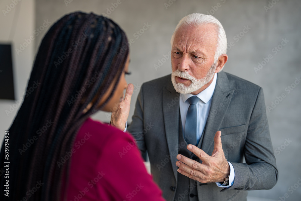 Angry senior executive manager in gray suit arguing with young female businesswoman blaming her for mistake at workplace. Upset senior businessman looking angry at sad overworked businesswoman. Angry senior executive manager in gray suit arguing with young female businesswoman blaming her for mistake at workplace. Upset senior businessman looking angry at sad overworked businesswoman.
