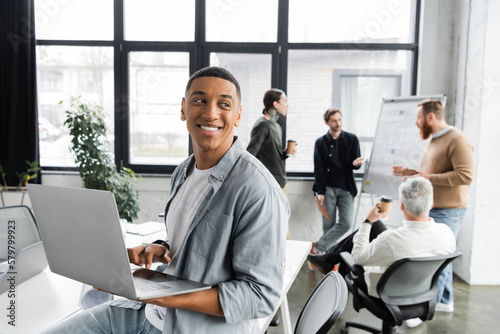Cheerful african american businessman using laptop near blurred colleagues working in office.