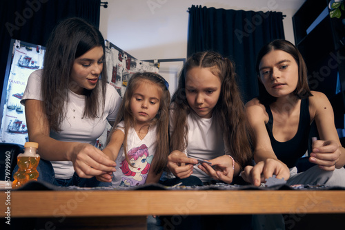 Staged photo. Lesbian couple and their kids are having a good time at home. Cozy atmosphere. Parents and girls are putting puzzles together. One of the parents helps the younger girl.