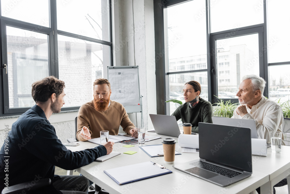 Bearded businessman talking to colleagues near devices and papers during training in office. Bearded businessman talking to colleagues near devices and papers during training in office.