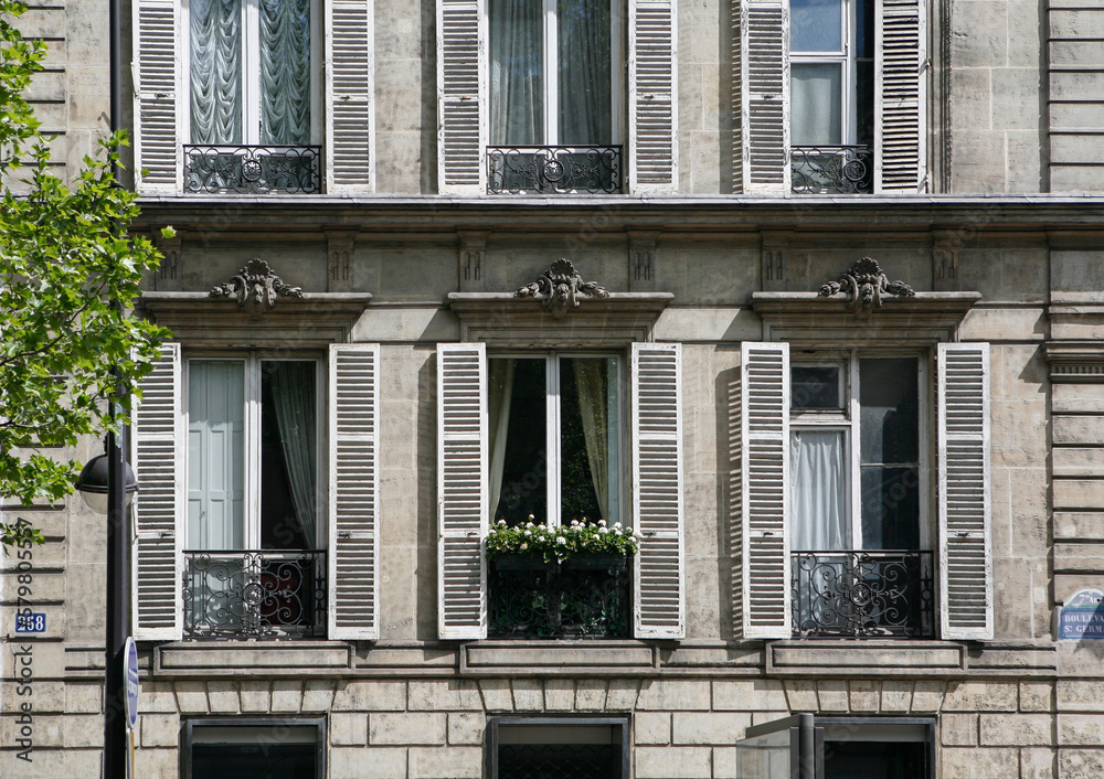 Obraz premium Paris ancient stone building facade with three rows of French windows with white wooden shutters, rich stucco fretwork, small wrought iron balconies, street lamp and tree branches on the left
