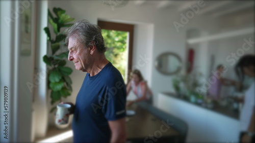 A contemplative senior man standing at home living room while drinking coffee. Mature older person staring out window