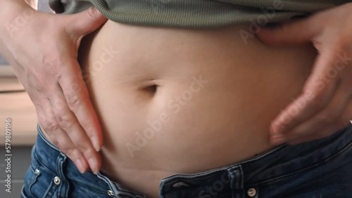 The woman pinches her belly fat disapprovingly with her fingers. White European girl in jeans and bare stomach.