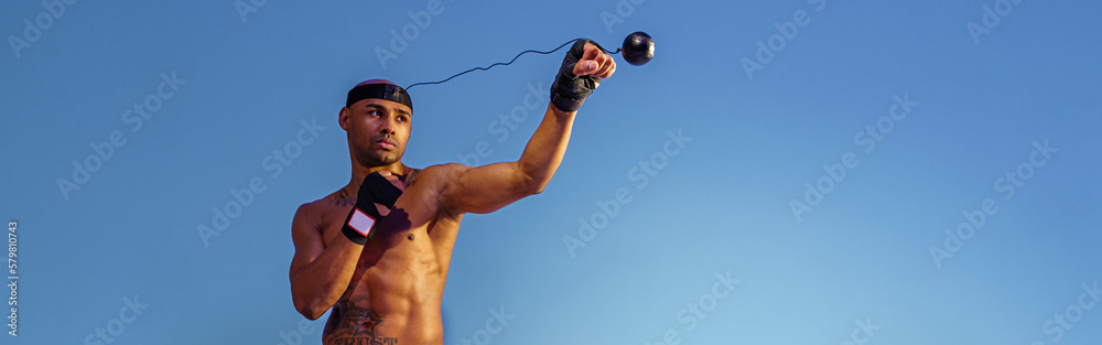 Sporty man with naked torso uses a boxing ball simulator for training on studio background