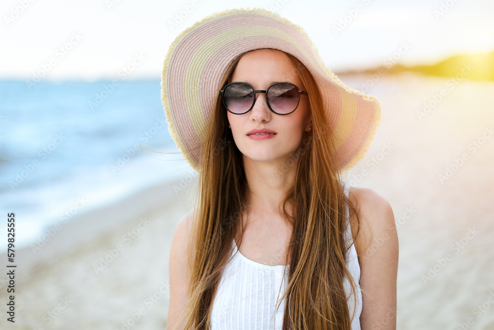 Portrait of a happy smiling woman in free happiness bliss on ocean beach standing with a hat and sunglasses. A female model in a white summer dress enjoying nature during travel holidays vacation