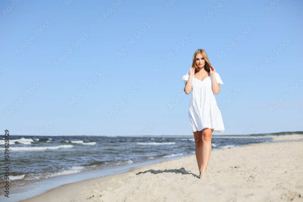 Happy, beautiful woman on the ocean beach standing in a white summer dress.