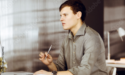 Young successful businessman in a green shirt is working on computer and is thinking about something, while sitting at the desk in a cabinet. Headshot or business portrait in an office.