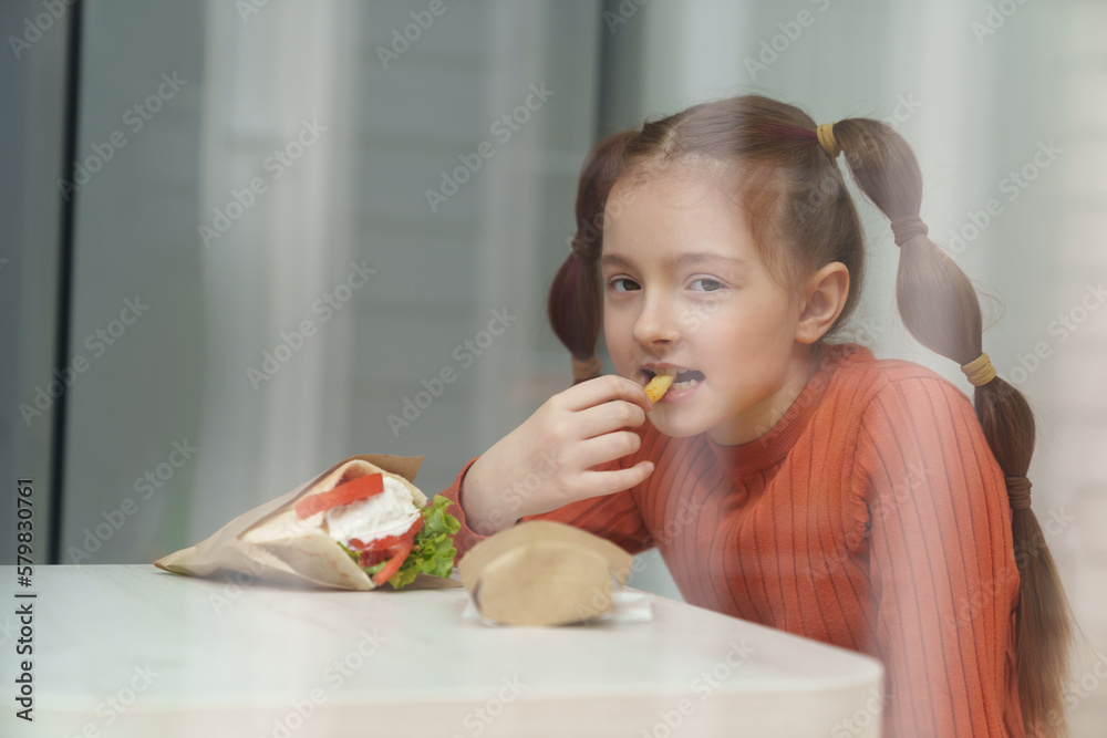 Cute little girl with ponytails eating souvlaki for lunch in Greek restaurant. Adroable white kid eats fries in Mediterranean cafe and looking in camera with a smile Cute little girl with ponytails eating souvlaki for lunch in Greek restaurant. Adroable white kid eats fries in Mediterranean cafe and looking in camera with a smile