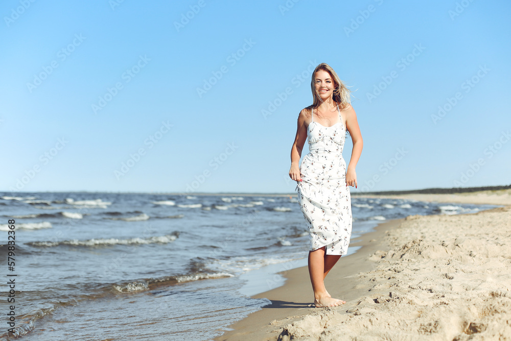 Happy blonde woman in free happiness bliss on ocean beach standing straight. Portrait of a female model in white summer dress enjoying nature during travel holidays vacation outdoors
