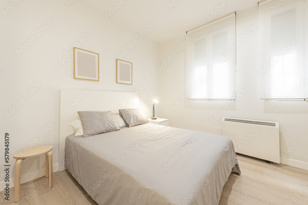 Fototapeta premium Bedroom with a double bed with wooden bedside tables, a gray bedspread with cushions, double windows with roller shutters and an electric heater